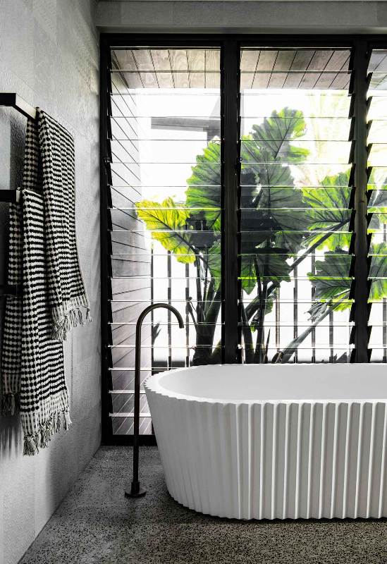 a contemporary tropical bathroom with grey marble tiles and terrazzo ones, a white sculptural bathtub, black fixtures and a glazed wall