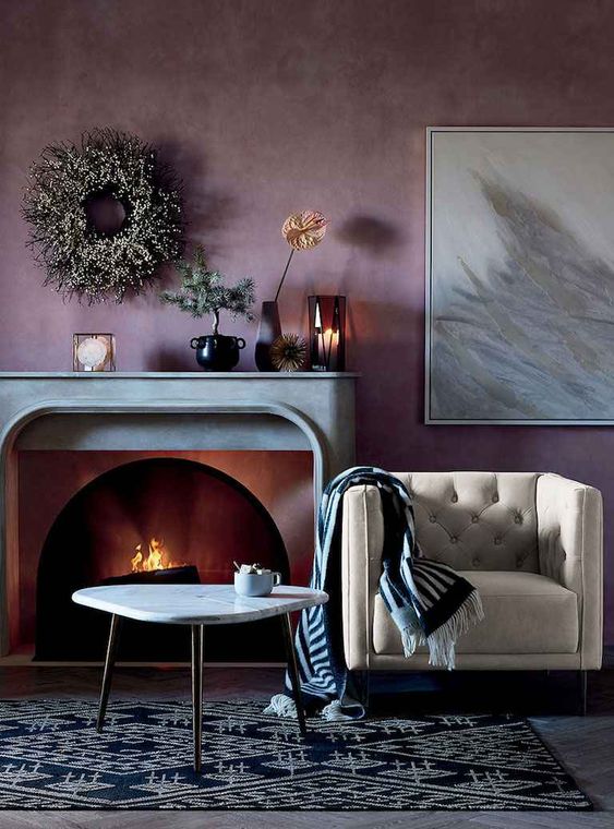 a dreamy living room with a pale purple accent wall, a fireplace with a stone mantel, a creamy chair, a printed rug and some lovely decor
