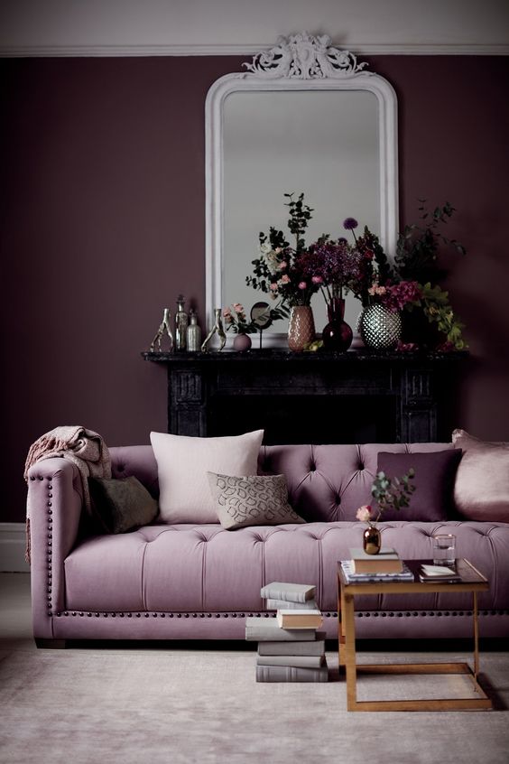 a gorgeous living room with a deep purple accent wall, a lilac sofa, a glass coffee table, a black fireplace and beautiful blooms on the mantel