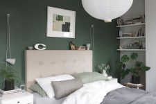 a lovely Scandinavian bedroom with a green accent wall, a tan upholstered bed, white nightstands and shelves, a pendant lamp and neutral bedding