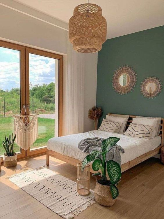 a lovely boho bedroom with a green accent wall, light stained furniture, a woven pendant lamp, printed textiles and sunburst mirrors plus some potted plants
