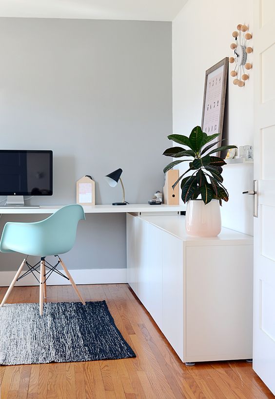 a lovely contemporary home office with a sleek storage unit and a built-in desk, a mint blue chair, potted greenery and a cool clock