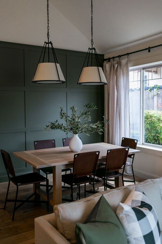 a modern dining space with a dark green paneled accent wall, a light stained table, brown leather chairs, pendant lamps and neutral curtains