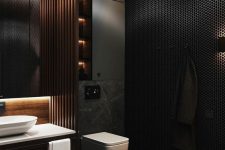 a moody contemporary bathroom with a black penny tile wall, a grey marble floor, a built-in vanity, a wooden beam screen and a lit up mirror