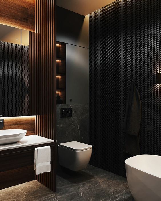 a moody contemporary bathroom with a black penny tile wall, a grey marble floor, a built-in vanity, a wooden beam screen and a lit up mirror