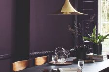 a moody dining room with a deep purple accent wall, a black trestle table and wooden chairs, a shiny gilded pendant lamp