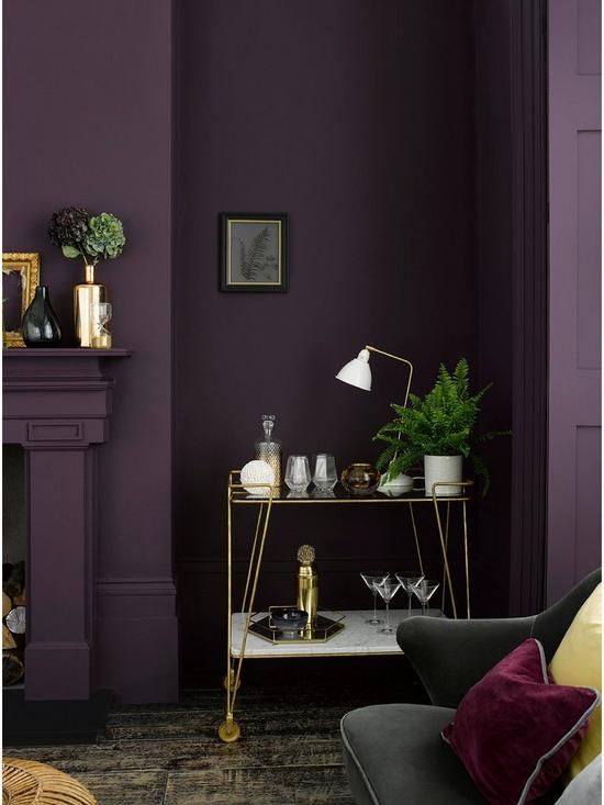 a moody living room with an aubergine accent wall, a fireplace, a delicate gold bar cart and a black sofa with colorful pillows