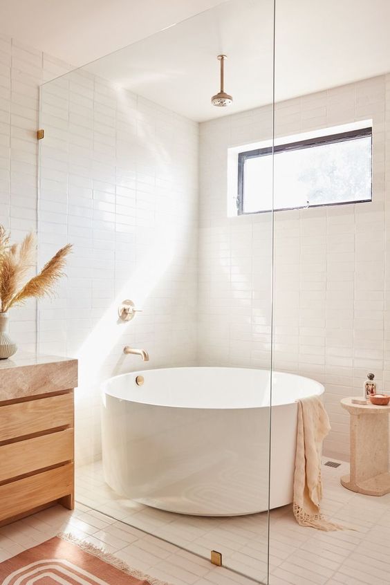 a neutral contemporary boho bathroom with white skinny tiles, a round tub, a wooden vanity and brass fixtures is a chic space