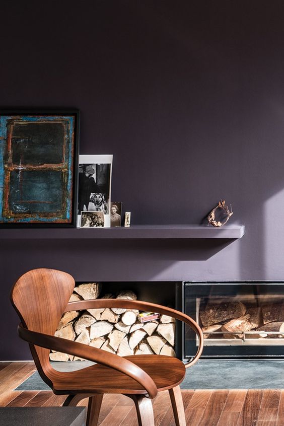a refined living room with a deep purple accent wall, firewood storage and a built-in fireplace, a floating shelf with decor