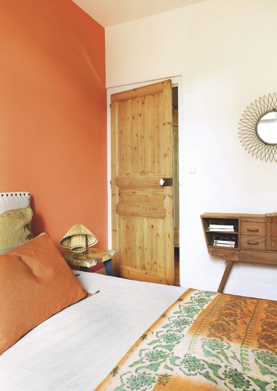 a small eclectic bedroom with a rust-colored accent wall, a vintage wooden sideboard and a cool nightstand with a woven lamp