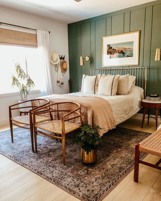 a stylish modern boho bedroom with a green paneled accent wall, a gilded bed, woven chairs and a bench, with printed bedding