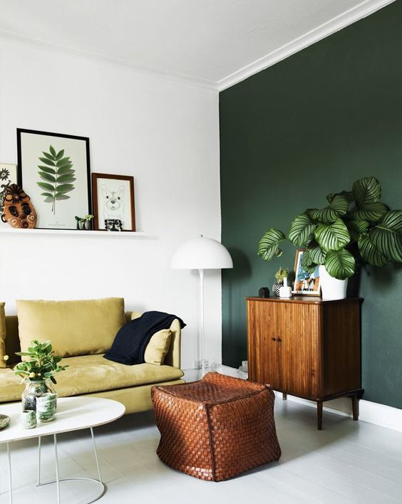 a stylish modern living room with a dark green accent wall, a mustard sofa, a brown leather pouf, potted greenery and a ledge gallery wall