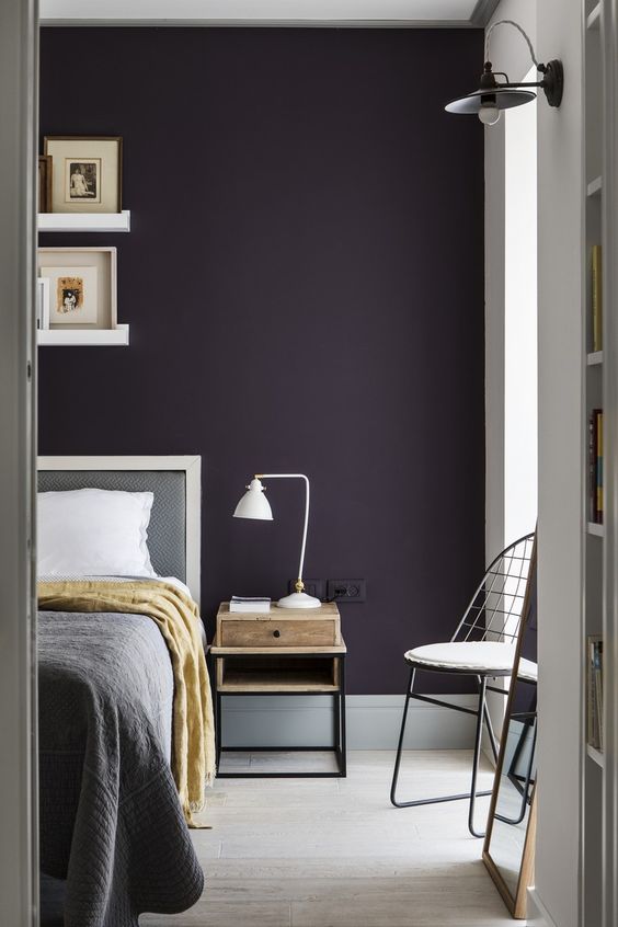 a stylish moody bedroom with a dark purple accent wall, grey and white furniture, a small nightstand with a table lamp and ledges with artworks