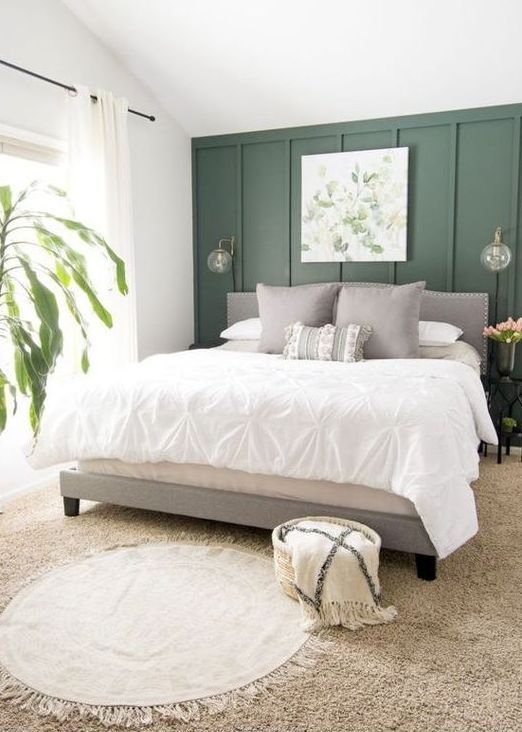 an organic bedorom with a green paneled accent wlal, a grey upholstered bed, neutral bedding, layered rugs and a botanical artwork and a potted plant