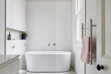 an ultra-minimalist bathroom with white and grey tiles, an oval tub, chrome fixtures and pastel textiles and a window for more light