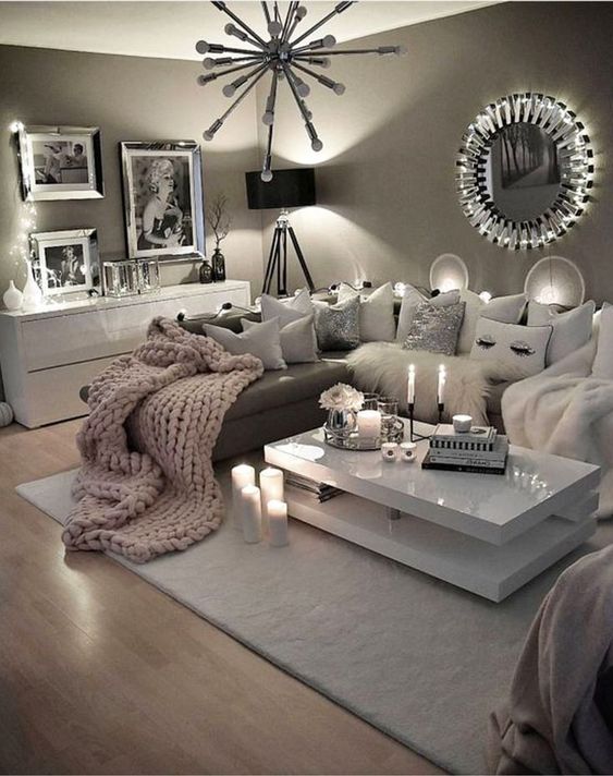a glam taupe living room with a brown sectional, a tiered white coffee table, a mini gallery wall, a white dresser and a sunbrust chandelier