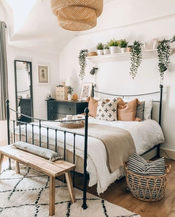 a stylish boho bedroom with a metal bed, a black dresser, a wooden bench, potted greenery, a woven lamp, a basket and a floor mirror