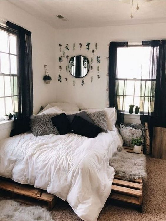 a lovely boho bedroom with white walls, a pallet bed, neutral bedding, grey and black pillows, a mirror with faux greenery and black curtains
