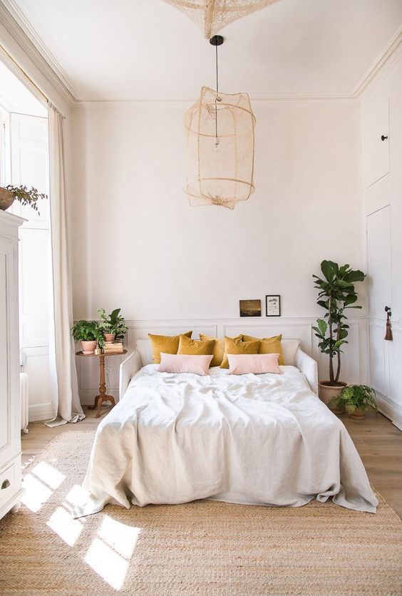 a pretty light-filled bedroom with paneled walls, a white bed, a pendant lamp, mustard and pink pillows and some potted greenery to refresh the space