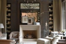 a sophisticated taupe living room with paneling, built-in bookshelves, a non-working fireplace, neutral seating furniture, a sphere pendant lamp and a large mirror