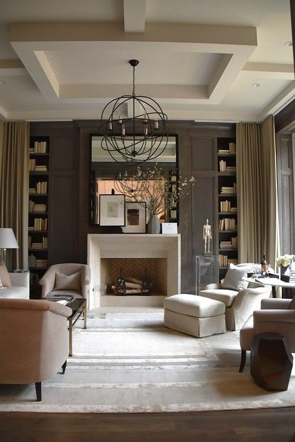 a sophisticated taupe living room with paneling, built-in bookshelves, a non-working fireplace, neutral seating furniture, a sphere pendant lamp and a large mirror