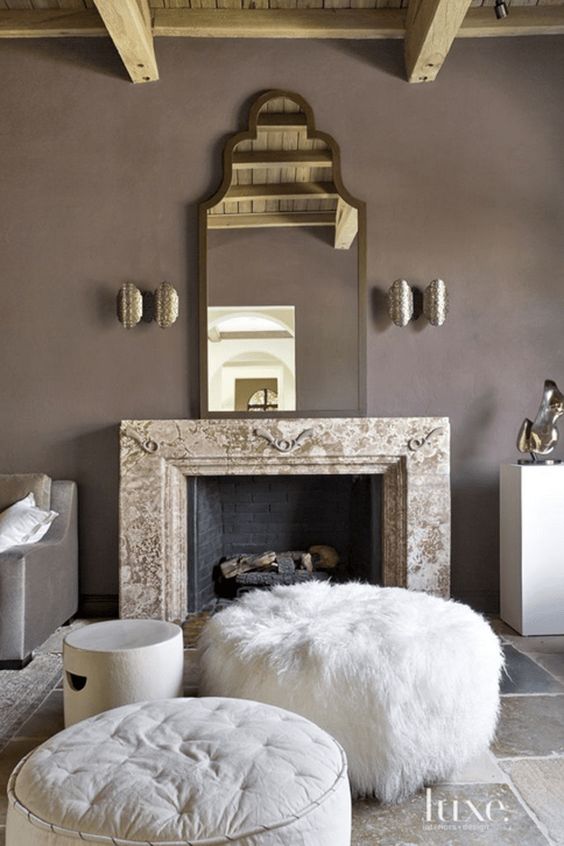 an exquisite taupe living room with a non-working fireplace, a refined mirror, metallic sconces, white ottomans and poufs and taupe furniture