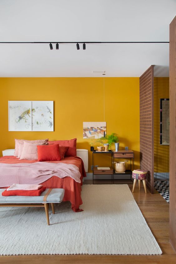 a chic bedroom with a mustard yellow accent wall, a white bed with colorful bedding, an upholstered bench and pretty artworks is cool