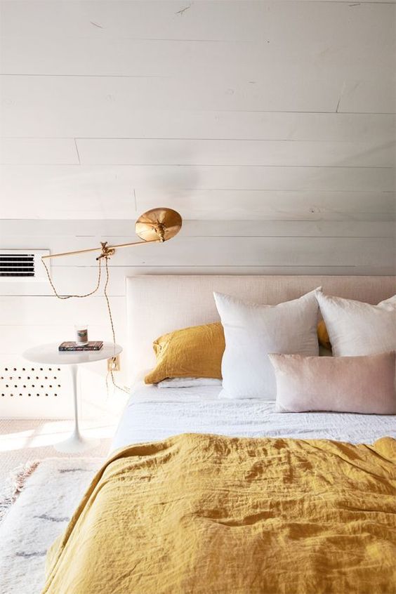 a dreamy and serene bedroom with white planked walls, a neutral upholstered bed, white and yellow bedding and a gold sconce is chic