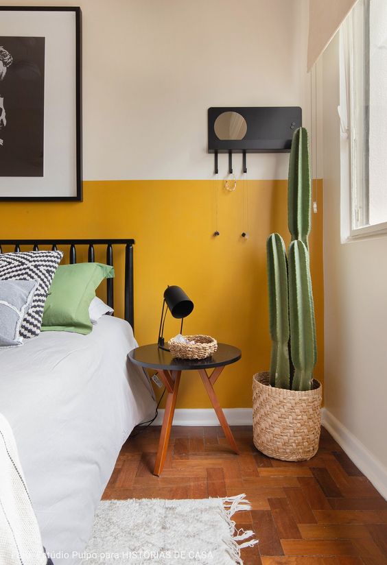 a modern boho bedroom with color block yellow and white walls, a blakc metal bed, a round table, a potted cactus and some art and pillows