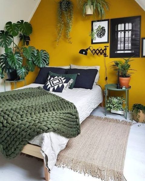 a pretty bedroom spruced up with Gen Z yellow - an accent wall, a bed with black, green and neutral bedding, potted plants, artworks and a green table is super cool
