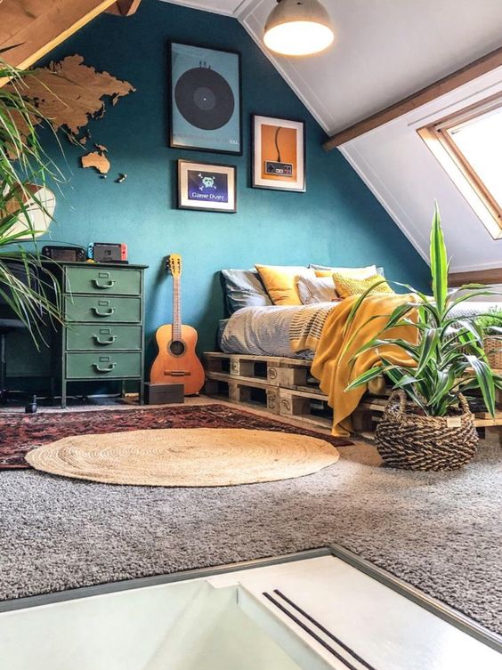 an attic bedroom with a pallet bed, yellow and grey bedding, a navy accent wall, a green dresser, a plywood map on the wall, layered rugs and potted plants