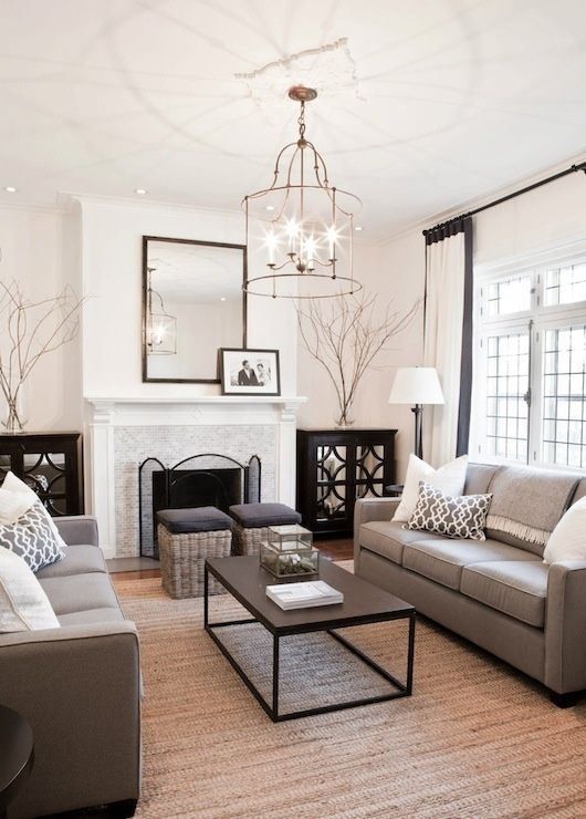 a traditional living room with taupe sofas, woven stools, creamy curtains and pillows, a chic chandelier and a non-working fireplace