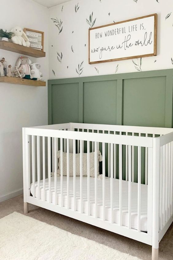 a cute and cozy nursery with green panels on the wall, a botanical wallpaper accent, a white crib, open shelves and neutral bedding