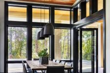 a lovely and simple contemporary dining room with a view of the forest, a simple stained table and black chairs, black pendant lamps