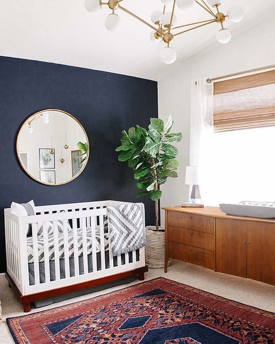 a mid-century modern nursery with a navy accent wall, a crib with printed bedding, a rich-stained changing table, a colorful printed rug and a chic chandelier