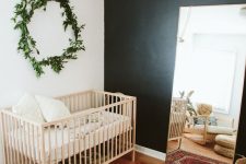 a modern boho nursery with a black accent wall, a stained crib and chair, a floor mirror, a printed rug, neutral bedding and a greenery wreath