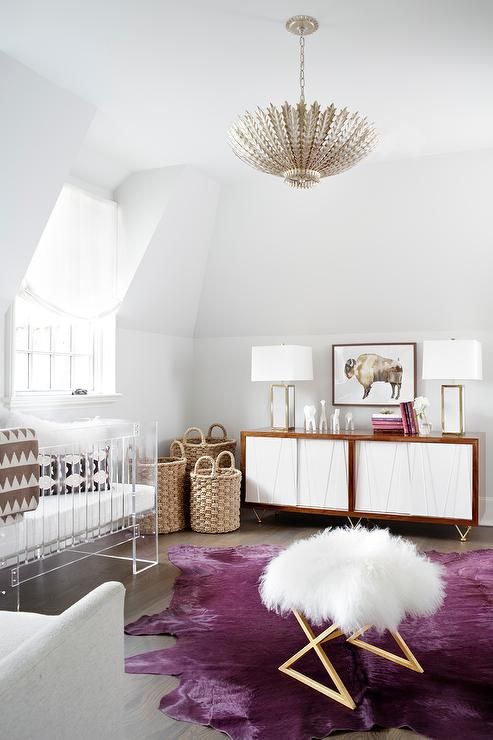 a modern glam nursery with an acrylic crib, a fluffy pouf, a white and stained credenza, baskets for storage and a purple statement rug