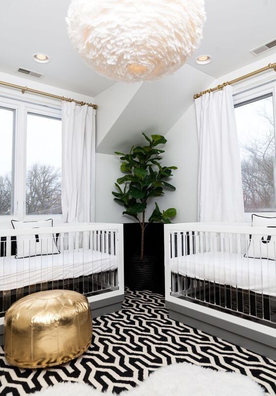 a modern twin nursery with a touch of glam, with acrylic cribs, a printed rug and bedding, a statement plant, a gold pouf and a pendant lamp