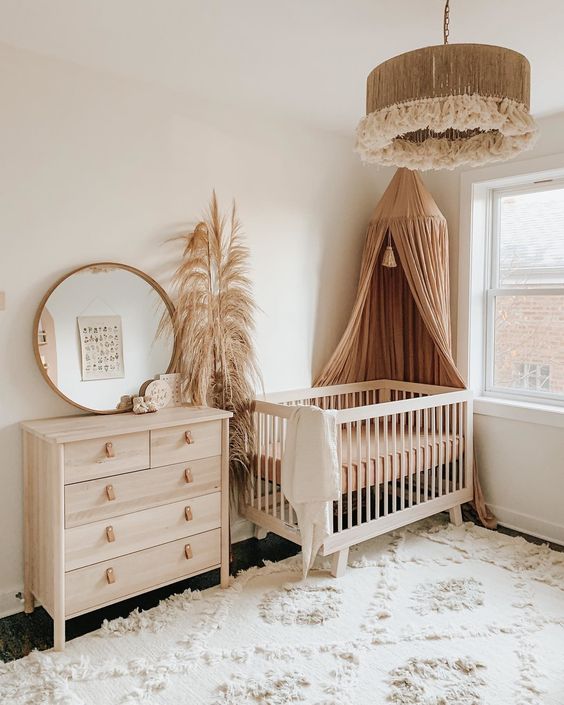 a neutral boho nursery with stained furniture, a rust-colored canopy, pampas grass, a round mirror and a cool tassel pendant lamp