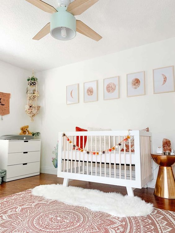 a pretty modern nursery with white furniture, moon prints, a lamp and a fan, a hanging shelf with toys, layered rugs and a shiny side table