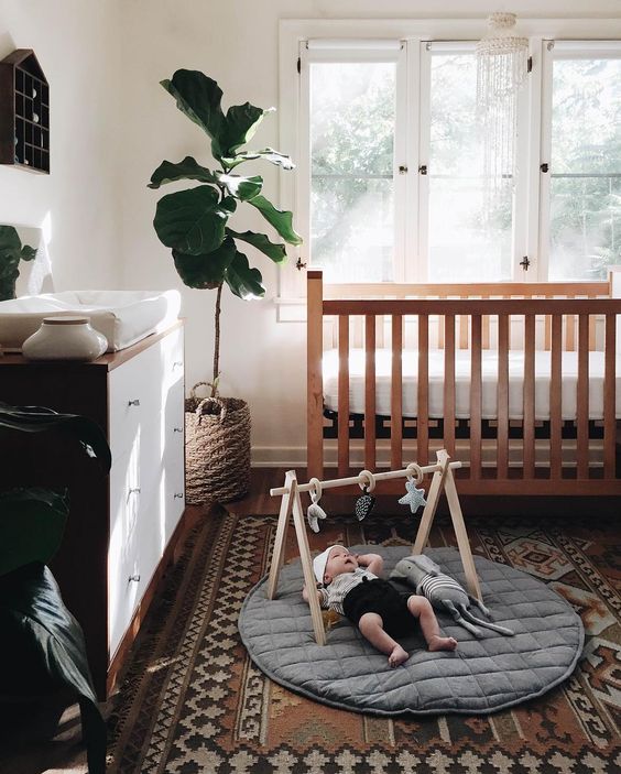 a stylish modern boho nursery with a stained crib, a stained and white dresser, a printed rug, a house-shaped shelf and statement plants