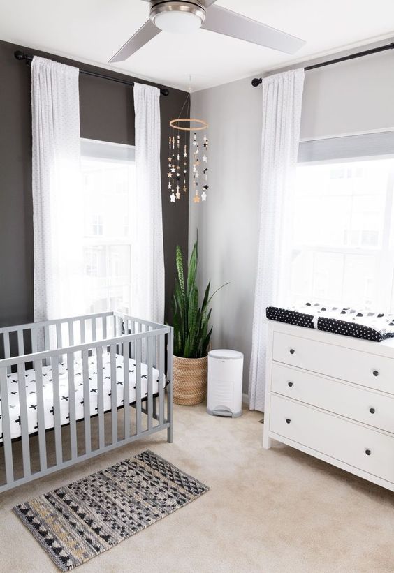 a stylish monochromatic nursery with a white dresser, a grey crib, a black accent wall, a printed rug and bedding, a mobile and white curtains