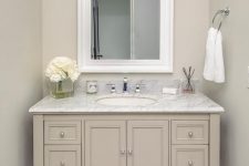 a tan bathroom with a tile floor, a taupe vanity with a white stone countertop, a mirror in a white frame and a sconce plus white blooms