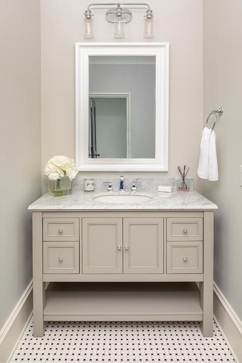 a tan bathroom with a tile floor, a taupe vanity with a white stone countertop, a mirror in a white frame and a sconce plus white blooms