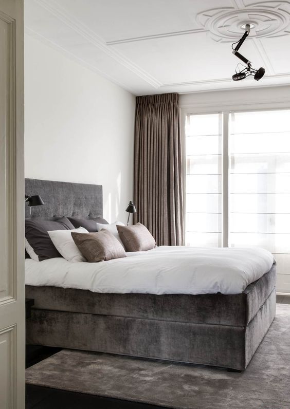 a white refined bedroom with a taupe upholstered bed, a matching rug, tan curtains and tan and taupe pillows