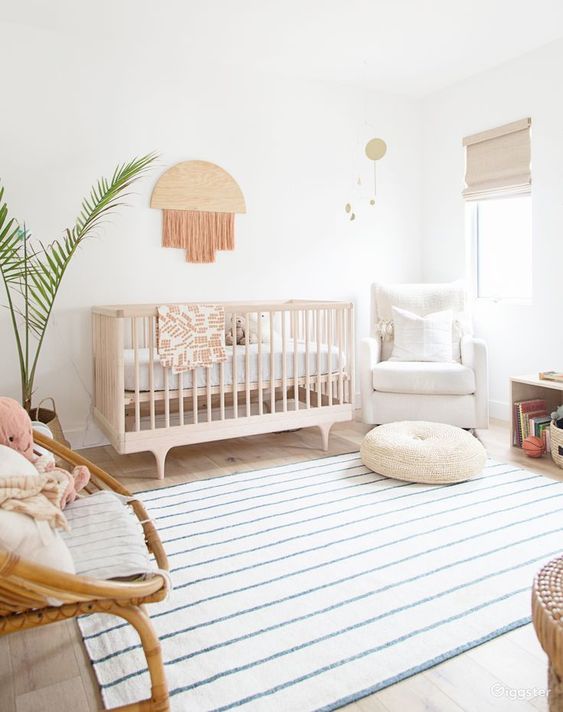 an airy boho nursery with a stained crib, a rattan and upohlstered chair, a fringe decoration, a printed rug plus a potted plant is a cool idea