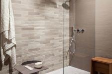 an elegant bathroom with taupe tiles on the walls, a chevron floor, neutral tiles for an accent wall, a taupe table and a wooden stool