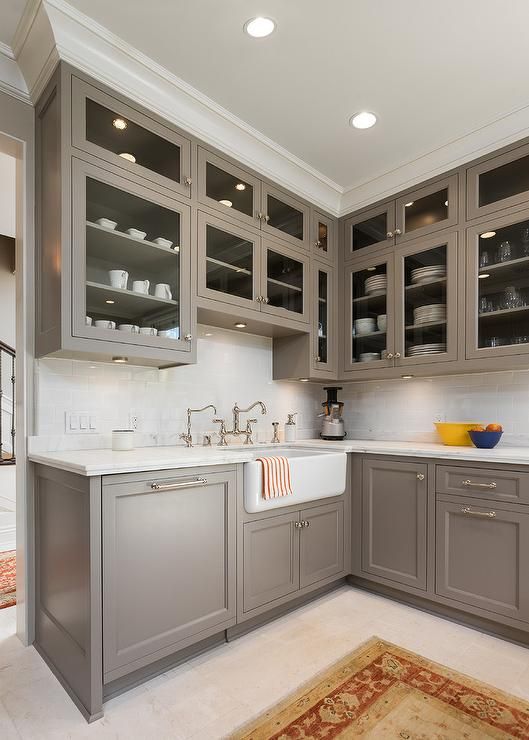 an elegant taupe vintage kitchen with shaker style cabinets, glass upper ones, a white stone countertop and a backsplash and vintage fixtures and knobs is all cool