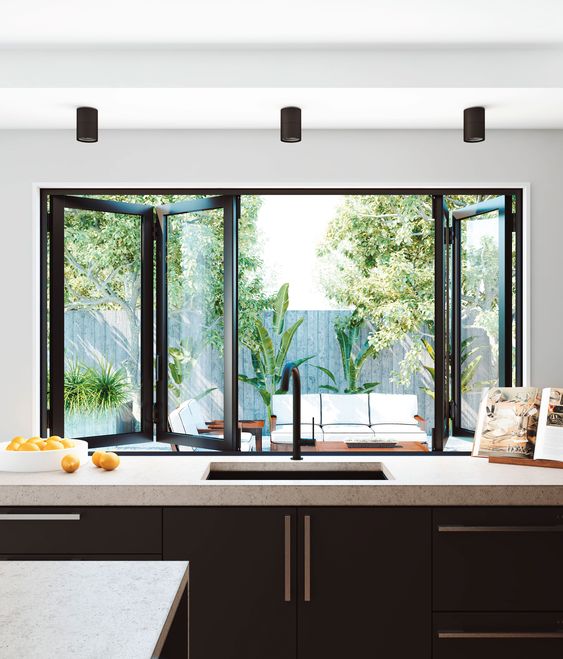 a black kitchen with neutral stone countertops, a black folding window with a view to the garden that can be opened any time