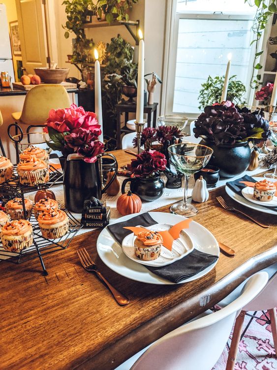 a Halloween tablescape with jugs with dark and bold blooms and a cauldron with dark flowers, candles and delicious cupcakes